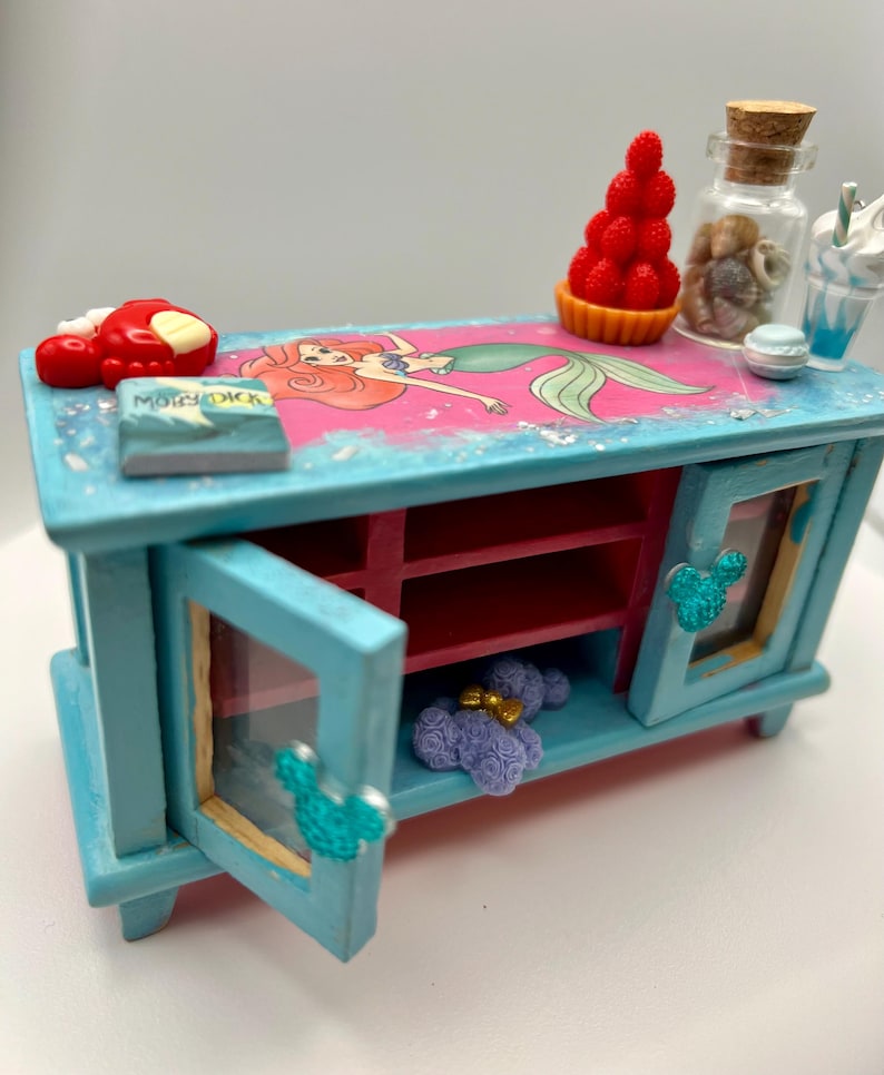May include: A miniature, hand-painted, turquoise cabinet with a pink and blue top featuring a mermaid illustration. The cabinet has two doors with Mickey Mouse-shaped handles, and is decorated with miniature food items.