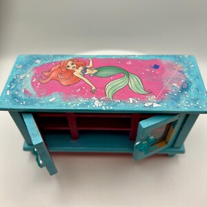 May include: A teal wooden jewelry box featuring a pink top with an illustration of a mermaid. The box has two small doors with a Mickey Mouse head design. The interior is red.