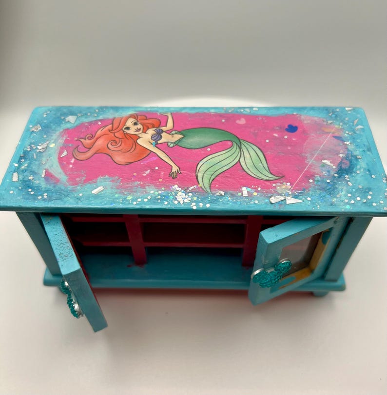 May include: A teal wooden jewelry box featuring a pink top with an illustration of a mermaid. The box has two small doors with a Mickey Mouse head design. The interior is red.