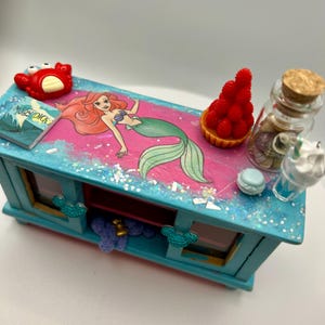 May include: A blue wooden jewelry box with a pink top featuring a mermaid illustration. The box is decorated with glitter and includes miniature accessories like a red crab, a book, a tart, a bottle with shells, and a drink.