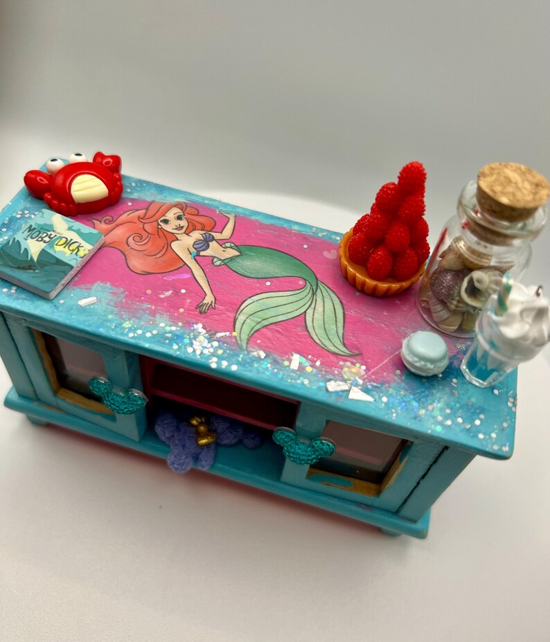 May include: A blue wooden jewelry box with a pink top featuring a mermaid illustration. The box is decorated with glitter and includes miniature accessories like a red crab, a book, a tart, a bottle with shells, and a drink.