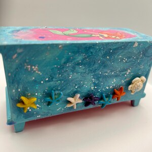 May include: A small, rectangular, blue wooden box with a mermaid illustration on the lid. The box is decorated with various starfish and a turtle. The box has a glittery, textured finish.