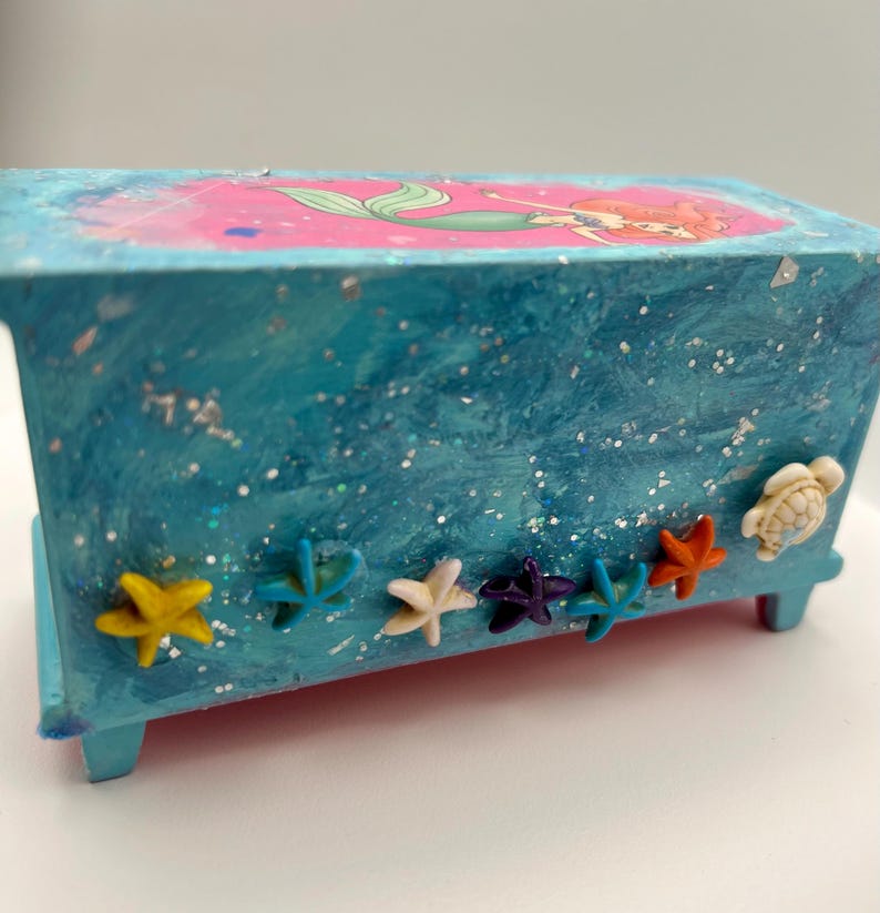 May include: A small, rectangular, blue wooden box with a mermaid illustration on the lid. The box is decorated with various starfish and a turtle. The box has a glittery, textured finish.
