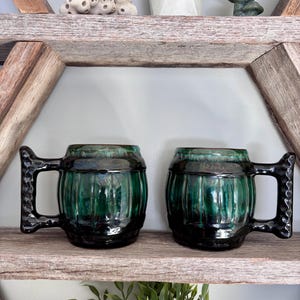 May include: Two ceramic mugs with a dark green glaze and black handles sit on a wooden shelf. The mugs have a ribbed design and a glossy finish. The shelf is part of a decorative wooden structure.