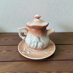 May include: A ceramic teapot and saucer set with a mushroom design. The teapot is a light brown colour with a white mushroom design and a matching lid. The saucer has a matching design. The set is on a wooden surface.