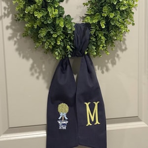 May include: A green wreath with a navy blue ribbon. The ribbon has an embroidered topiary design and the letter "M" in yellow. The wreath is made of artificial greenery and is hanging on a door.