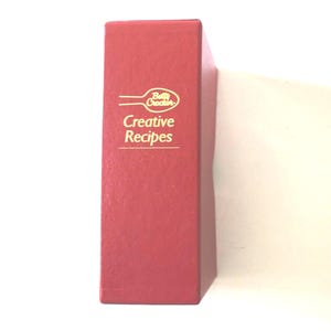 May include: A red cookbook titled "Creative Recipes" by Betty Crocker. The book has gold lettering and a gold logo. The book is standing upright and is on a white surface.