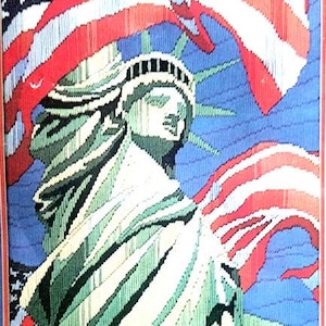 May include: Needlepoint artwork depicting the Statue of Liberty with the American flag in the background. The statue is green and white, set against a blue sky. The flag features red and white stripes and white stars.