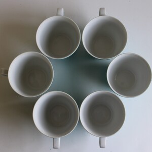 Pu&ograve; includere: Sei tazze in ceramica bianca con bordi blu, disposte in cerchio su una superficie bianca.