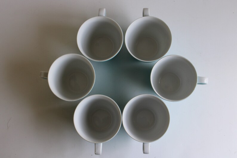 Pu&ograve; includere: Sei tazze in ceramica bianca con bordi blu, disposte in cerchio su una superficie bianca.