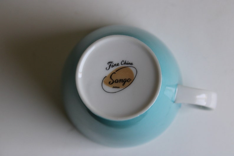 Pu&ograve; includere: Una tazza da t&egrave; blu chiaro con interno bianco e un logo dorato e nero che recita "Fine China Sango Japan".