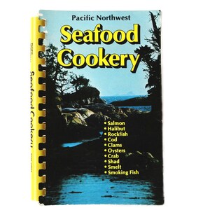 May include: A yellow and black spiral bound cookbook titled "Pacific Northwest Seafood Cookery". The cover features a scenic photo of a rocky coastline with a blue sky and water. The book lists various types of seafood including salmon, halibut, rockfish, cod, clams, oysters, crab, shad, smelt, and smoking fish.