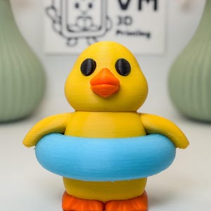 May include: A 3D-printed yellow duck figurine with a blue inner tube. The duck has black eyes and an orange beak and feet. The background includes a sign with the text "VM 3D Printing".