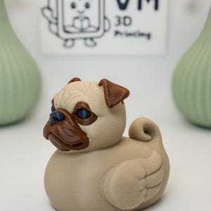 May include: A 3D-printed pug figurine in a duck shape. The pug has a tan body, brown ears and snout, and blue eyes. The wings are detailed with feather-like lines. The background includes a logo and two green vases.