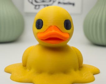 May include: A yellow 3D printed duck figurine with a bright orange beak and dark eyes. The base of the duck is flattened and spread out, creating a melted appearance. In the background, a white sign displays the text VM 3D Printing.