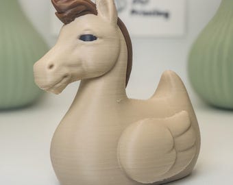 May include: A tan-colored, 3D-printed hybrid figurine of a horse and a duck. The figurine has a horse's head with a brown mane and a duck's body with small wings. The background includes a sign with the text "VM 3D Printing".