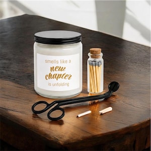 Puede incluir: Una vela blanca en un frasco de vidrio transparente con tapa negra, con una etiqueta que dice "smells like a new chapter is unfolding" en letras doradas. Junto a ella hay un frasco pequeño con cerillas y un cortamechas de metal negro.