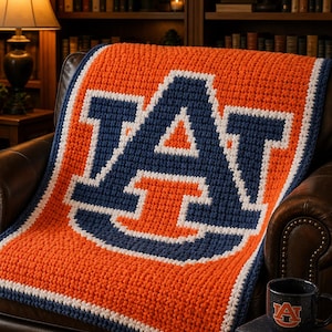 May include: A crocheted blanket in orange and navy blue, featuring a large white-outlined letter "A". The blanket is draped over a brown leather armchair. A matching mug and books are on a side table.