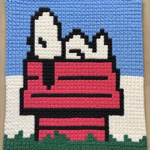 Può includere: Una coperta quadrata all'uncinetto fatta a mano con un design pixelato di Snoopy. La coperta mostra Snoopy nella sua cuccia rossa contro un cielo blu, con accenti bianchi e verdi. La coperta è realizzata con filato morbido.