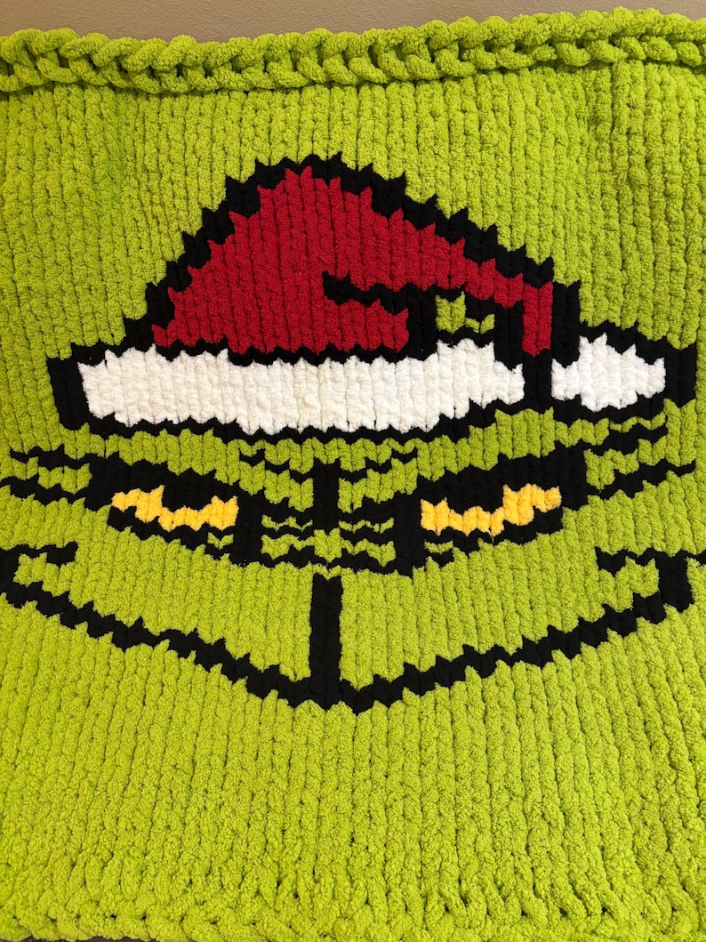 May include: A lime green, pixelated blanket featuring a Christmas character design. The design includes a red Santa hat with a white trim, black outlines, and yellow eyes. The blanket is made of a soft, chunky knit material.