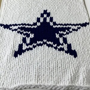 Op de afbeelding: Een wit en marineblauw gebreid deken. De deken heeft een grote marineblauwe ster in het midden, omgeven door een witte achtergrond. De deken is gemaakt van een zacht, dik gebreid materiaal en de randen zijn netjes afgewerkt.