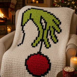 May include: A cream-colored crocheted blanket featuring a pixelated design of a green hand holding a red ornament with a black outline. The blanket is draped over a beige armchair, with a Christmas tree and fireplace in the background.
