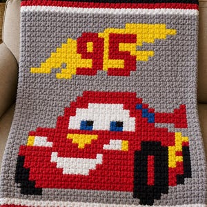 May include: A handmade crochet blanket featuring a red race car design with the number 95 and yellow flames. The blanket has a gray background with black, white, and red borders. The blanket is displayed on a beige sofa.
