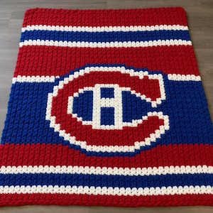 May include: A handmade blanket in red, white, and blue. The blanket features a large emblem of a white "H" inside a red "C", with white and blue stripes. The blanket is made of a thick, textured yarn. The blanket's dimensions are in centimetres.