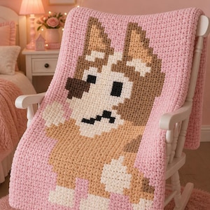 Puede incluir: Una manta rosa tejida a ganchillo con un diseño de perro de dibujos animados en tonos marrón, beige y blanco. La manta está sobre una mecedora blanca. El diseño es pixelado. La manta añade calidez y confort.