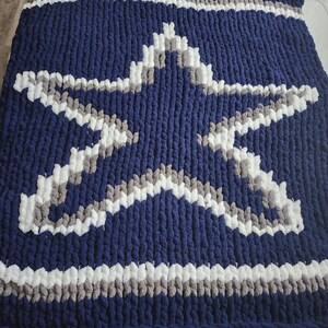 Op de afbeelding: Een marineblauwe, dik gebreide deken met een groot sterontwerp in wit en grijs. De deken heeft witte en grijze strepen langs de boven- en onderranden. Deze gezellige deken is perfect om comfort toe te voegen aan elke kamer.