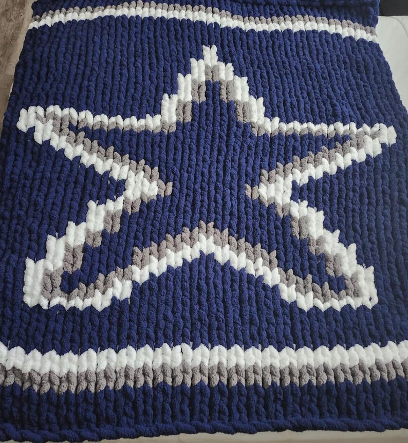 Op de afbeelding: Een marineblauwe, dik gebreide deken met een groot sterontwerp in wit en grijs. De deken heeft witte en grijze strepen langs de boven- en onderranden. Deze gezellige deken is perfect om comfort toe te voegen aan elke kamer.