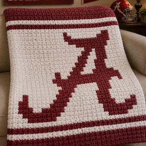 May include: A handmade crochet blanket in crimson and white, featuring the letter "A" in the center. The blanket has a textured appearance with stripes and a solid border. The words "ROLL TIDE" are visible in the background.