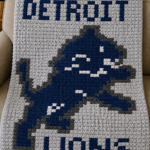 Könnte beinhalten: Eine gehäkelte Decke in Grau mit den Worten "DETROIT LIONS" in marineblauen Blockbuchstaben. Die Decke zeigt auch ein pixeliges Löwenlogo in Marineblau und Grau. Die Decke ist über ein beiges Sofa drapiert.