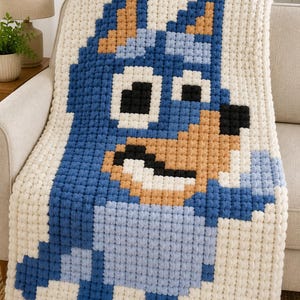 Può includere: Una coperta fatta a mano, in stile pixelato, raffigurante un personaggio di cane blu su sfondo crema. La coperta è realizzata con filato morbido e spesso, con il viso e il corpo del cane in tonalità di blu, beige e nero. La coperta è drappeggiata su un divano beige.
