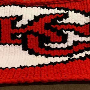May include: A hand-knitted red, white, and black blanket featuring the Kansas City Chiefs logo. The logo is an arrowhead shape with the letters "KC" in red and black, set against a white background. The blanket is made of thick yarn.