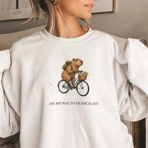 May include: White sweatshirt featuring a whimsical illustration of a capybara riding a bicycle with a basket of flowers. The text "ON MY WAY TO DE-ESCALATE" is printed below the image. A cozy, casual top.