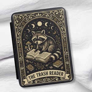 May include: Black and gold rectangular case featuring a raccoon reading a book, with the text "THE TRASH READER" on a banner. The design includes moons, stars, and mushrooms. The case has a black border.