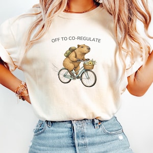 May include: Cream-colored t-shirt with the text "OFF TO CO-REGULATE". The shirt features an illustration of a capybara riding a light blue bicycle with a basket of flowers. The capybara wears a backpack.