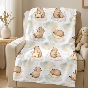 May include: A white baby blanket with a repeating pattern of cartoon capybaras, clouds, stars, and moons. The capybaras are in various poses, some holding stars, others cuddling. The blanket is draped over a beige armchair.