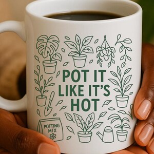 May include: White ceramic mug with the phrase "POT IT LIKE IT'S HOT" in green, surrounded by plant illustrations. The mug is filled with dark liquid and held in hands.