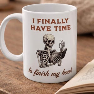 Puede incluir: Taza de cerámica blanca con un esqueleto negro y marrón leyendo un libro. El texto "I FINALLY HAVE TIME to finish my book" es marrón. Un libro abierto está sobre una superficie de madera.