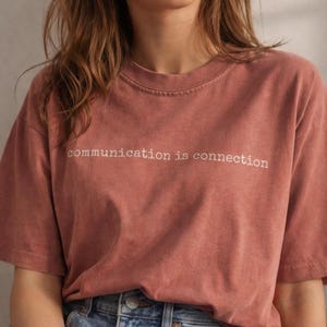 May include: A dusty rose-colored t-shirt with the phrase "communication is connection" printed in white. The shirt has short sleeves and a relaxed fit. The model is wearing blue jeans.