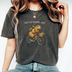 May include: A charcoal gray short-sleeve t-shirt featuring a graphic of a capybara riding a light blue bicycle. The capybara wears a green backpack and has a basket of flowers on the handlebars. The text OFF TO MODEL AAC is printed above.
