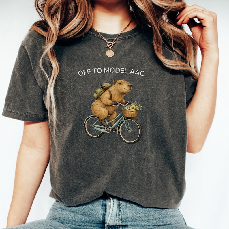 May include: A charcoal gray short-sleeve t-shirt featuring a graphic of a capybara riding a light blue bicycle. The capybara wears a green backpack and has a basket of flowers on the handlebars. The text OFF TO MODEL AAC is printed above.
