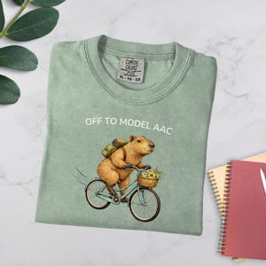 May include: A sage green t-shirt featuring a graphic of a brown capybara riding a light blue bicycle. The capybara wears a green backpack and carries a basket of yellow flowers. The text above the graphic reads OFF TO MODEL AAC.