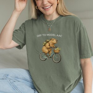 May include: Olive green t-shirt with the text "OFF TO MODEL AAC." The shirt features a whimsical illustration of a capybara riding a bicycle with a basket of flowers. The capybara wears a backpack.