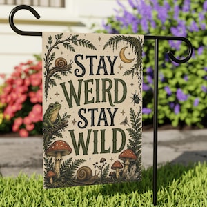 May include: A garden flag with the words "STAY WEIRD STAY WILD" in dark green lettering. The flag features illustrations of mushrooms, snails, a frog, and a crescent moon, all set against a beige background. The flag is displayed on a black metal stand.