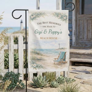 May include: A rectangular garden flag with a beach scene and the text "THE BEST MEMORIES ARE MADE AT Gigi & Pappy's BEACH HOUSE." The flag features a lighthouse, beach chair, and floral accents. The flag is displayed on a black metal stand.