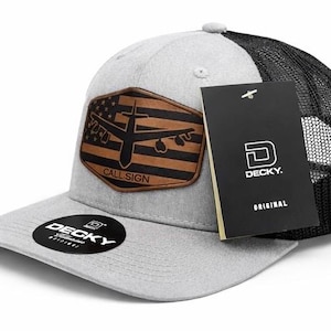 May include: A gray and black trucker hat with a brown leather patch featuring an airplane silhouette over a flag design. The patch reads "CALL SIGN." The hat has a curved brim and a "DECKY" tag.