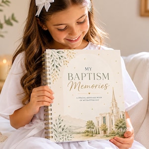 Peut inclure: Un livre à spirales intitulé "MY BAPTISM Memories" avec une illustration à l'aquarelle d'une église et d'un paysage. La couverture présente des lettres dorées et des accents botaniques. Le livre est tenu par une personne portant une robe blanche.